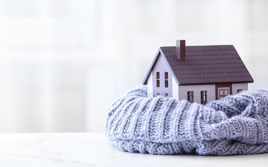 Miniatura de uma casa protegida por um cobertor de lã cinza, simbolizando conforto térmico no lar. Imagem ilustrativa para post sobre como deixar a casa mais quentinha no inverno, do blog da Soma Urbanismo.