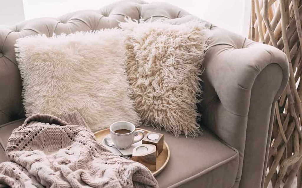 Sofá com almofadas felpudas, manta de tricô e bandeja com xícara de café e velas, sugerindo conforto e bem-estar.