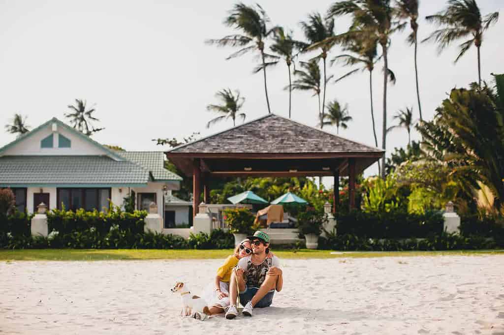 Foto de um casal abraçado e um cachorro sentados na areia e uma casa de verão ao fundo, para ilustrar o texto sobre casa de verão no blog da Soma Urbanismo. 