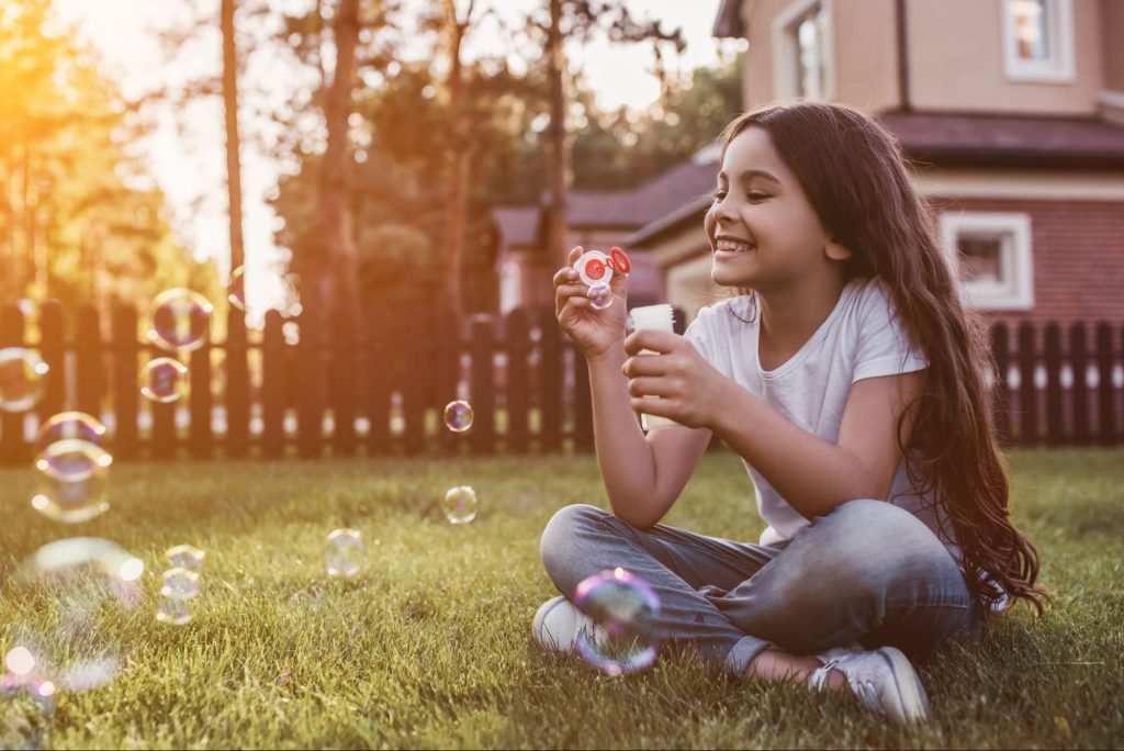 Foto de menina sentada na grama fazendo bolhas de sabão para ilustrar o texto sobre brincar com as crianças no quintal. 