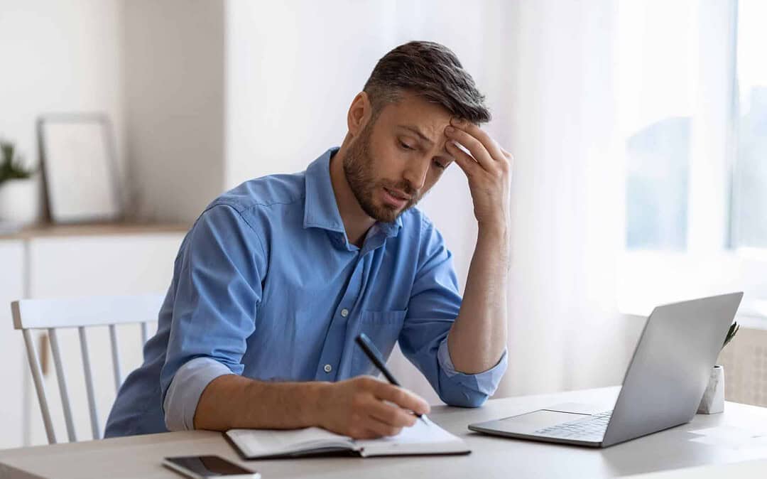 De camisa azul e em frente ao notebook, homem, preocupado, faz as contas e calcula investimento na construção de sua casa. Imagem ilustrativa para post "Construindo sua casa: a importância do cronograma para reduzir custos".