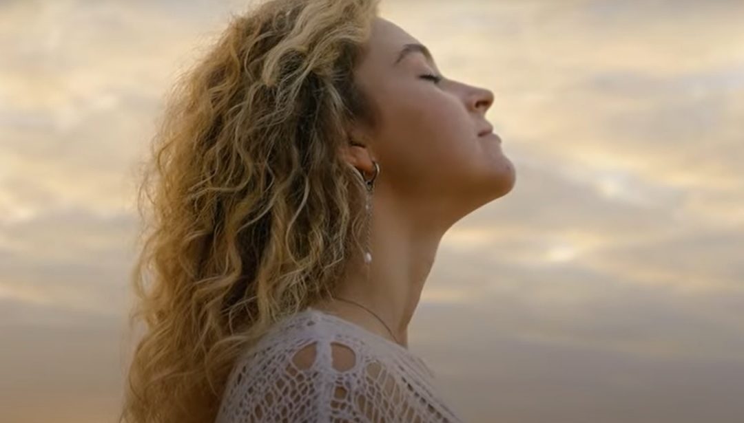 Mulher de perfil com os olhos fechados, expressão serena e cabelos ao vento, apreciando o momento ao ar livre sob um céu suave ao entardecer.