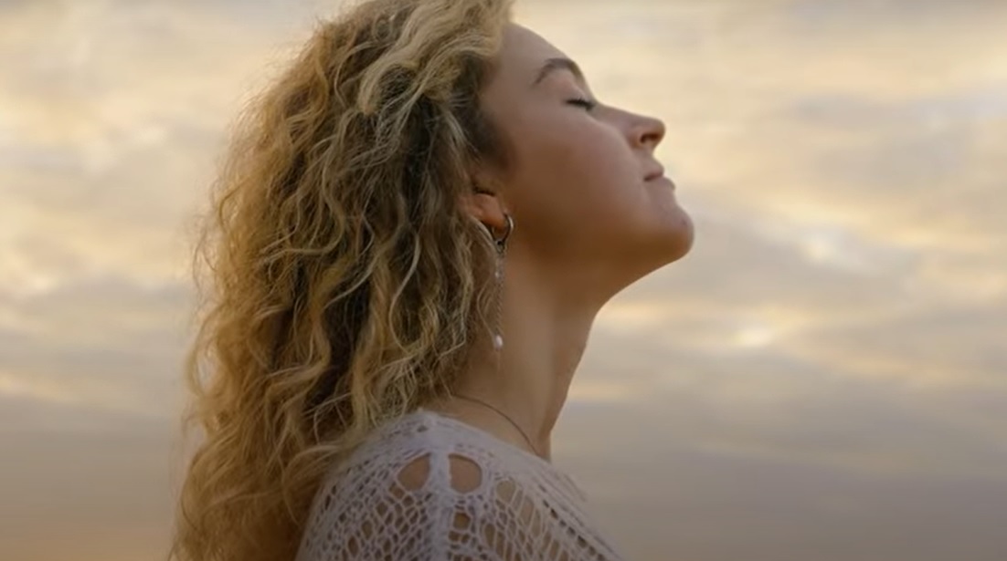 Mulher de perfil com os olhos fechados, expressão serena e cabelos ao vento, apreciando o momento ao ar livre sob um céu suave ao entardecer.