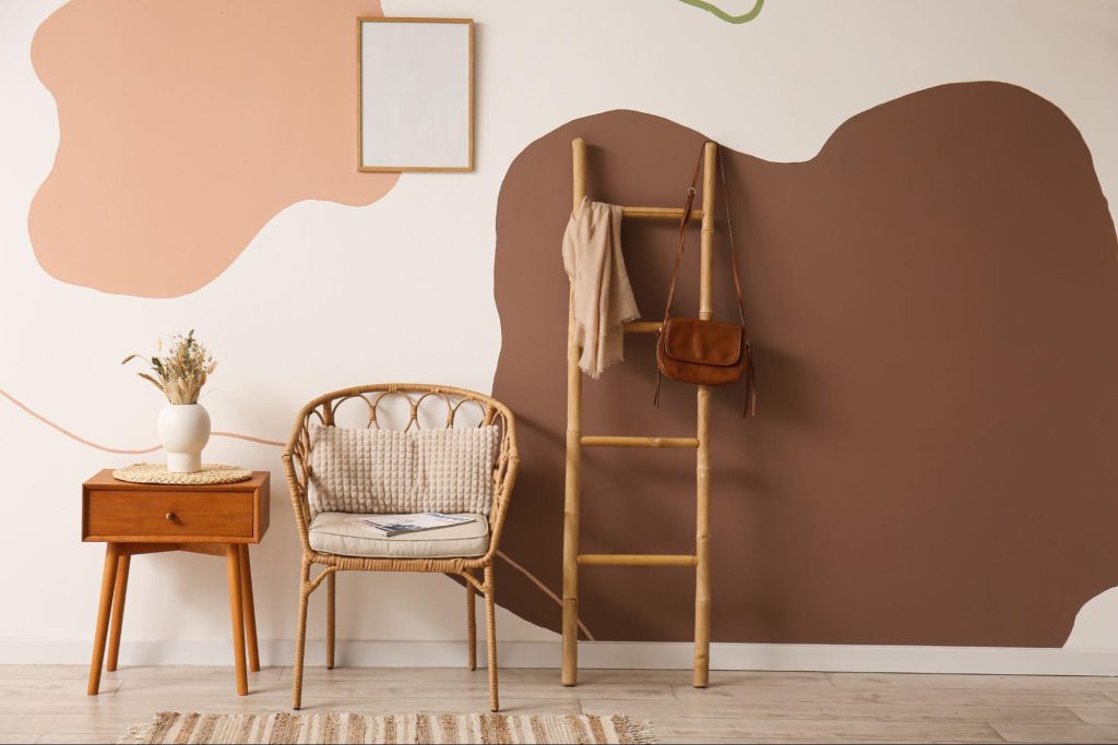 Ambiente decorado com parede pintada em formas orgânicas terrosas, cadeira de vime com almofada bege, mesa lateral com vaso de flores secas, e escada de bambu com bolsa marrom e echarpe penduradas. Imagem ilustrativa para post sobre decoração de outono, do blog da Soma Urbanismo.