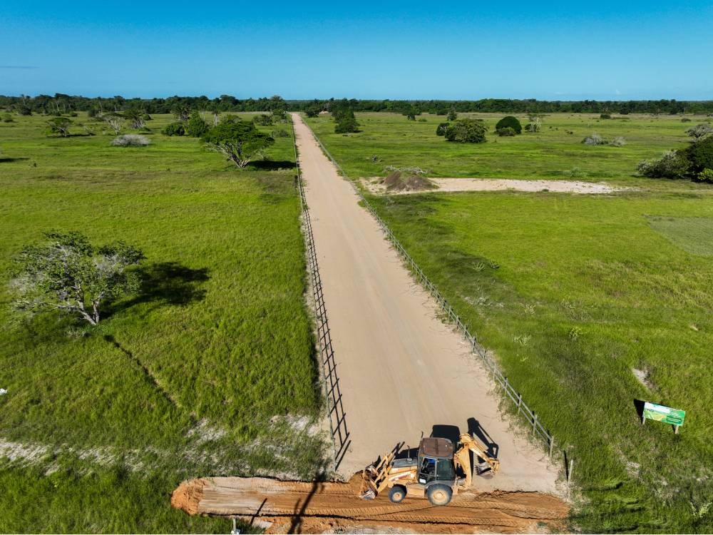 Trator trabalhando na abertura de estrada de terra em meio a uma ampla área verde, com vegetação rasteira e árvores esparsas sob céu azul claro, simbolizando o início de um nova gleba rural em desenvolvimento. Imagem do Vista Verde Espaços Rurais, da Soma Urbanismo.