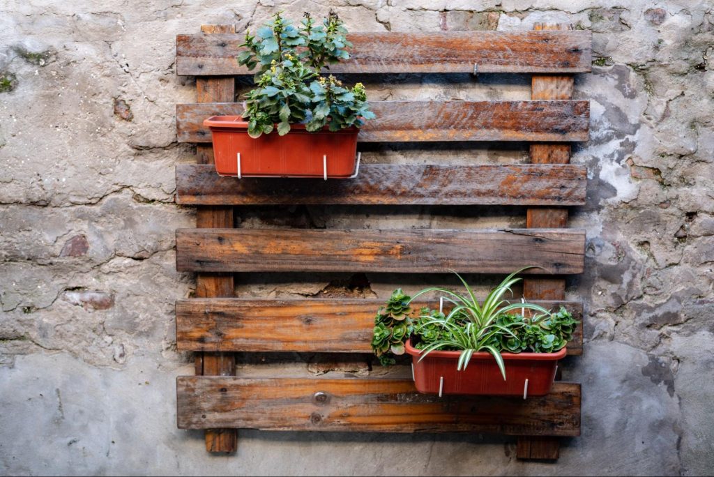 Jardim vertical rústico com floreiras presas a um pallet de madeira em parede de pedra. Imagem ilustrativa para post sobre modelos de jardim de inverno, do blog da Soma Urbanismo.