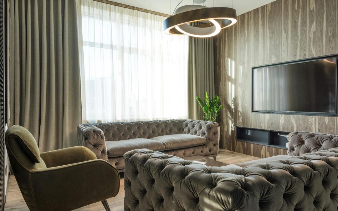 Sala de estar moderna com sofá capitonê cinza, poltrona verde, parede com revestimento amadeirado, TV de tela plana, cortinas longas e luminária de design contemporâneo. Imagem ilustrativa para post sobre tendências de decoração ultrapassadas, do blog da Soma Urbanismo.