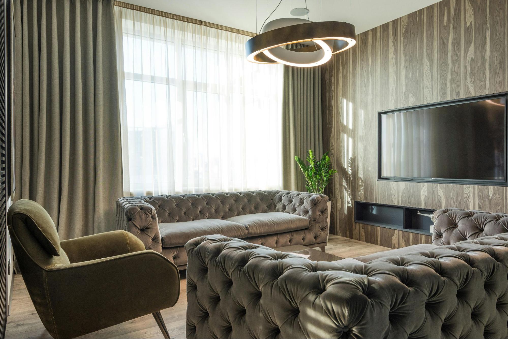 Sala de estar moderna com sofá capitonê cinza, poltrona verde, parede com revestimento amadeirado, TV de tela plana, cortinas longas e luminária de design contemporâneo. Imagem ilustrativa para post sobre tendências de decoração ultrapassadas, do blog da Soma Urbanismo.