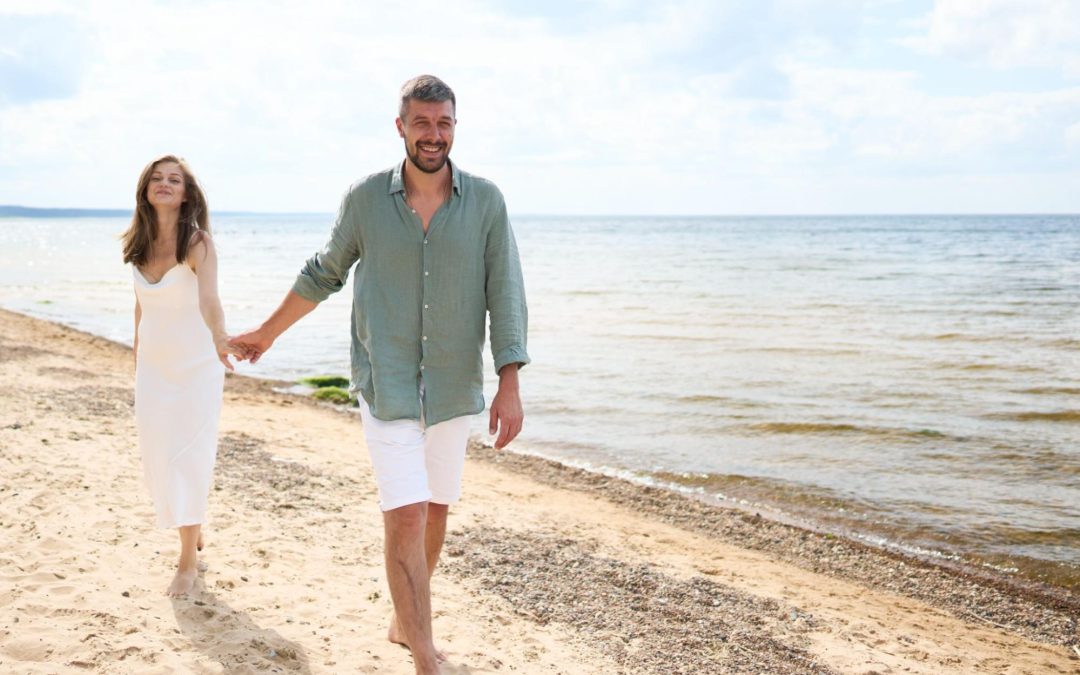 Casal caminha de mãos dadas pela faixa de areia de uma praia, com o mar ao fundo, representando qualidade de vida, bem-estar e o estilo de vida proporcionado por imóveis no litoral capixaba.