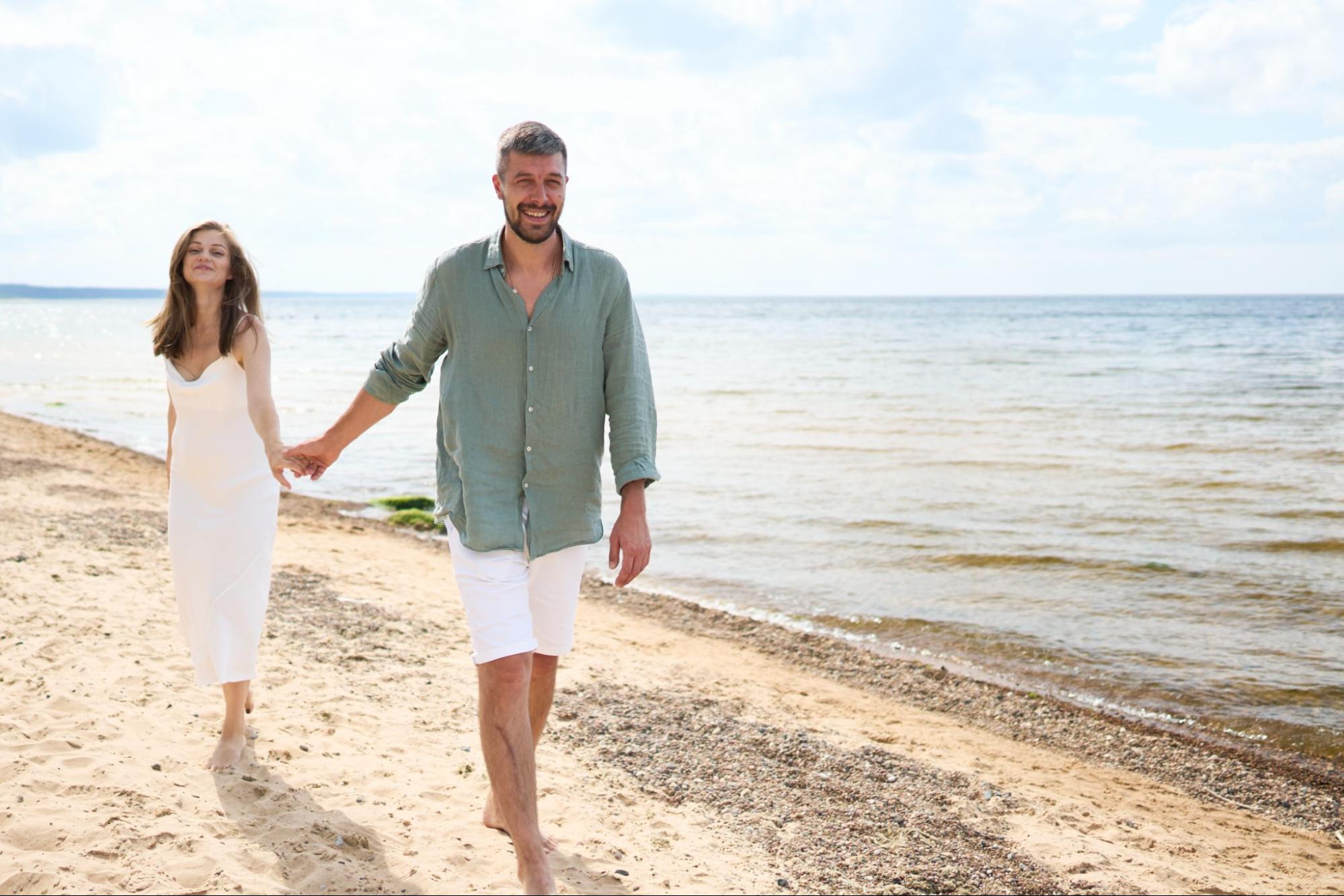 Casal caminha de mãos dadas pela faixa de areia de uma praia, com o mar ao fundo, representando qualidade de vida, bem-estar e o estilo de vida proporcionado por imóveis no litoral capixaba.