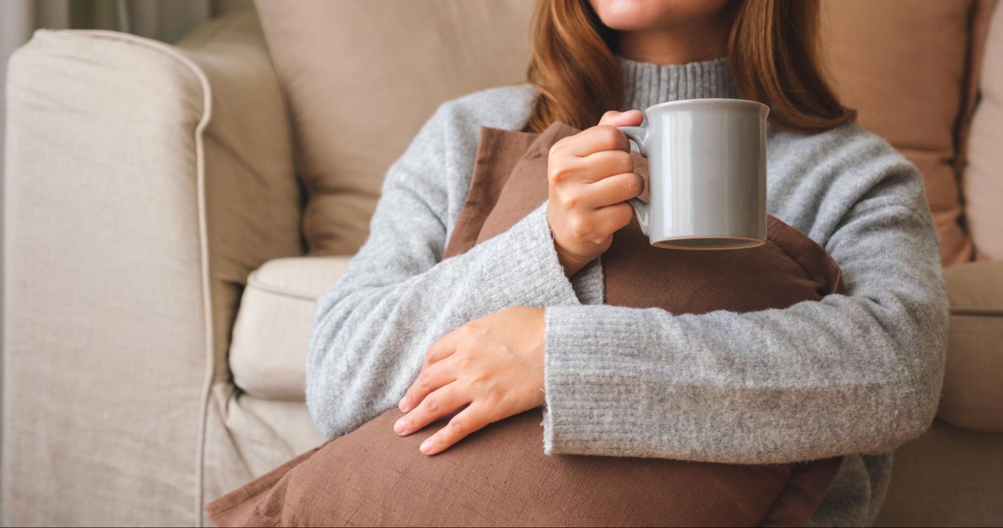 Mulher com casaco sentada em sofá e tomando algo quente em caneca. Imagem ilustrativa para post sobre o quanto a qualidade de vida de um bairro pode ser impactada durante o inverno.